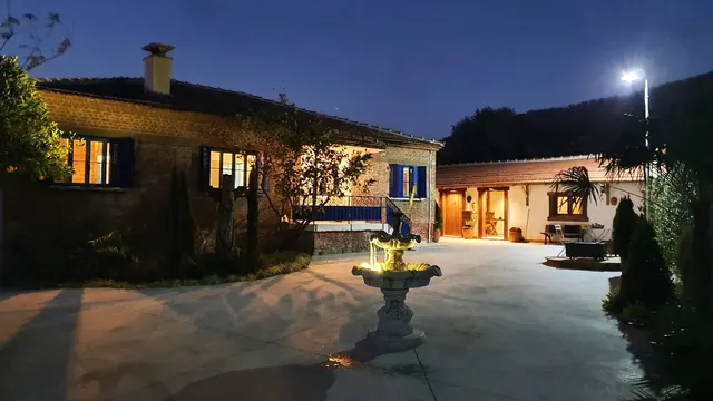 Akşam saatlerinde aydınlatılmış avlu; ortada ışıklandırılmış taş çeşme, solda mavi pencereli tuğla bina, sağda kiremit çatılı ek yapı.